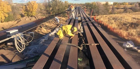 Abrasive media blasting—worker in protective gear using blasting equipment to clean and resurface steel beams on an outdoor structure.