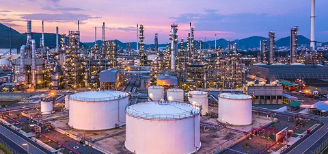 Large chemical plant facility with storage tanks, towers, and pipelines illuminated at dusk with mountains in the background.