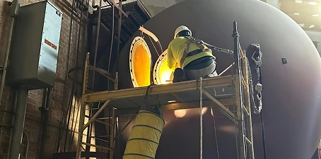 Worker in protective gear on scaffolding applying coating inside a large confined industrial tank opening, lit by work lights.