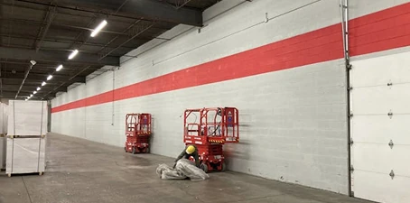 Workers using lifts to paint a long warehouse interior wall with a bold red stripe across gray concrete.