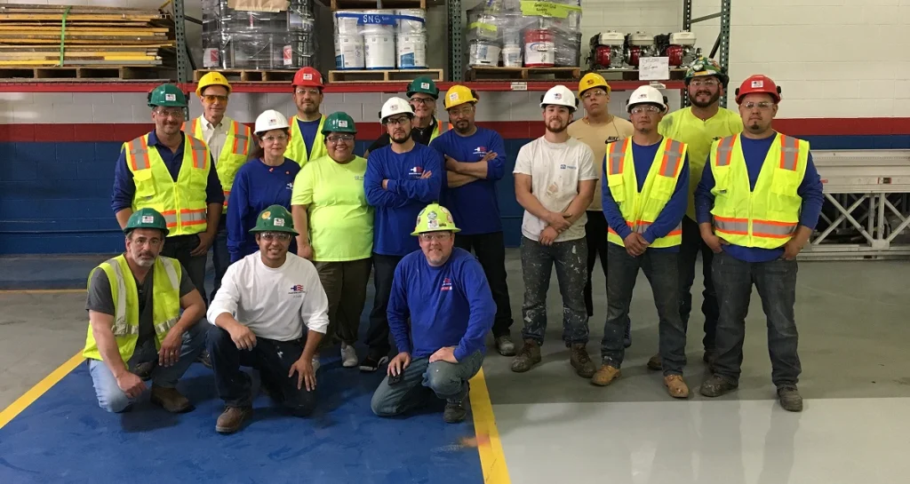 A group of sixteen construction and industrial workers pose for a team photo inside a warehouse or workshop. They are wearing safety gear including hard hats, safety vests, and work boots. The group is diverse, with both men and women standing and kneeling on a clean, painted concrete floor. Behind them are shelves stocked with equipment, tools, and supplies. The mood appears friendly and professional.