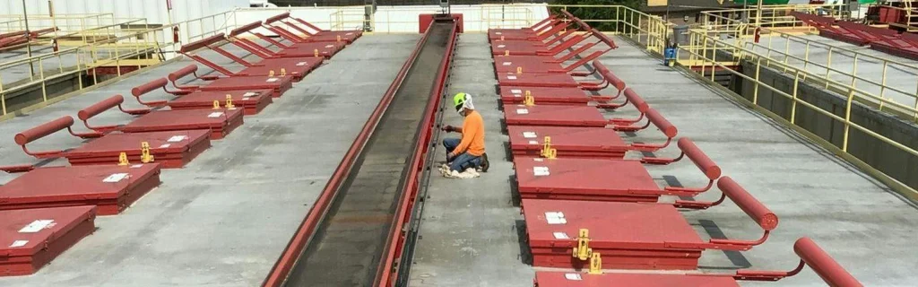 Worker in safety gear kneels on an industrial roof between long rows of propped-open red hatches and guardrails.