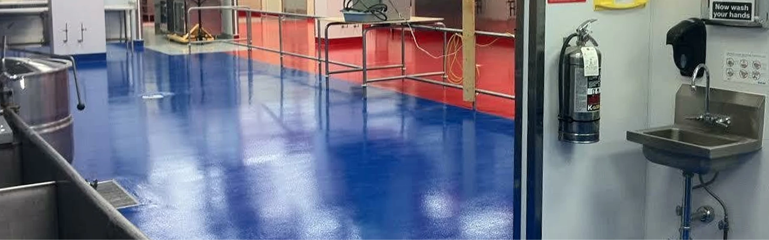 Clean industrial food processing area with shiny blue epoxy floor, stainless steel equipment, a wall-mounted handwashing sink, and a fire extinguisher near the entrance.
