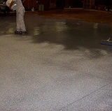 Worker applying a non-slip flooring finish with a roller, leaving a textured, slip-resistant surface across the concrete floor.