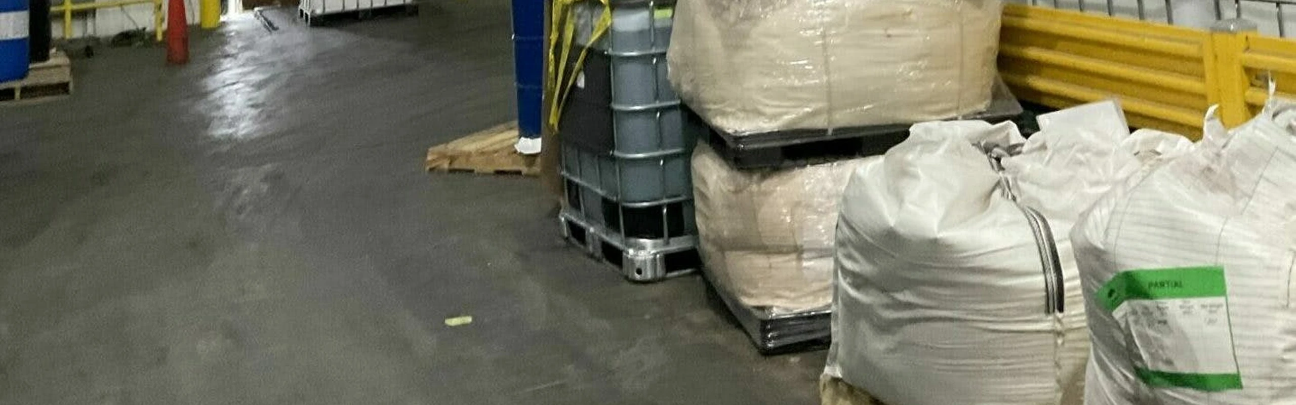 Warehouse aisle with dark concrete floor; stacked bulk bags and a plastic IBC tote on pallets sit beside blue drums and yellow safety guardrails.