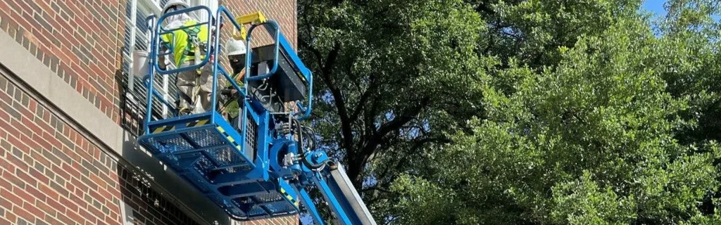 Workers in safety gear on a blue lift painting the brick exterior of a multi-story building next to large trees.