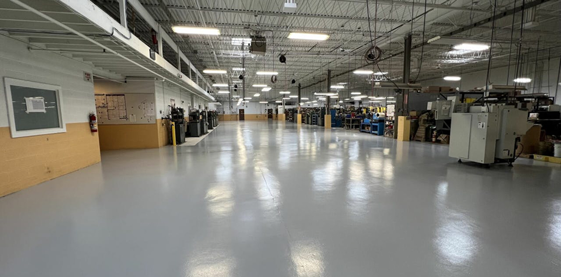 Large manufacturing floor with a smooth, glossy gray coating under bright overhead lights; equipment and workstations line the perimeter of the spacious, clean facility.