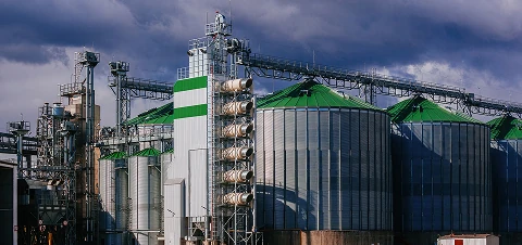 Large grain facility with multiple metal silos and green-roofed storage tanks connected by overhead conveyors under a cloudy sky.