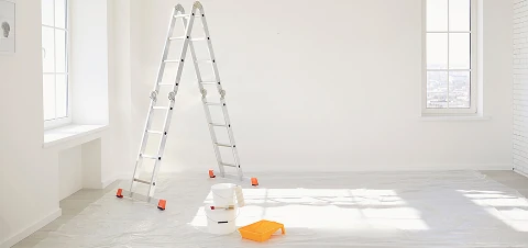 Bright empty room prepared for painting, with an aluminum ladder, paint bucket, and roller tray on a plastic-covered floor near tall windows.